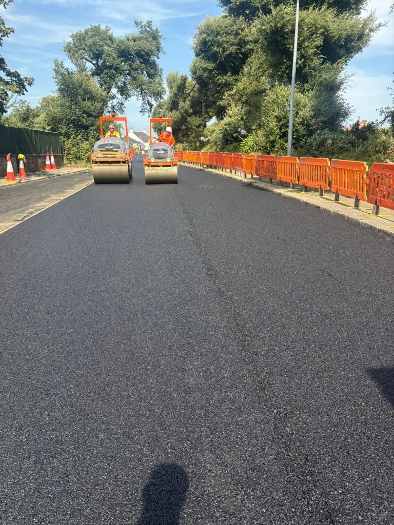 Two road rollers compacting freshly laid asphalt on a public carriageway with safety barriers