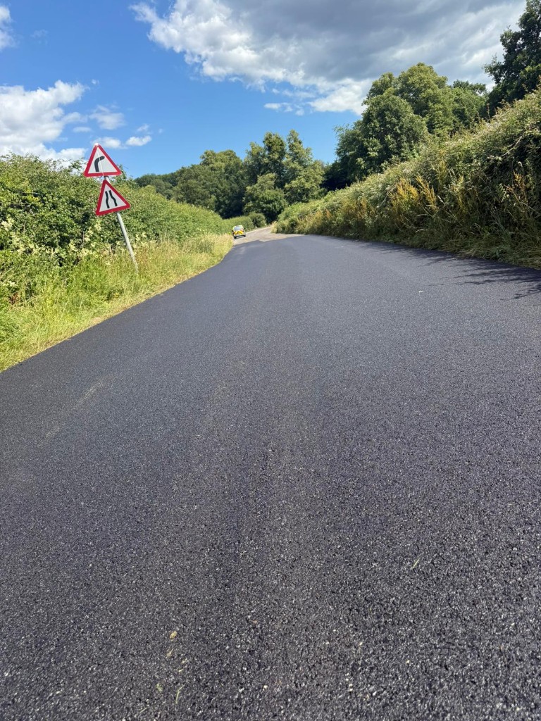 Freshly resurfaced rural road bordered by hedges with advance warning signage