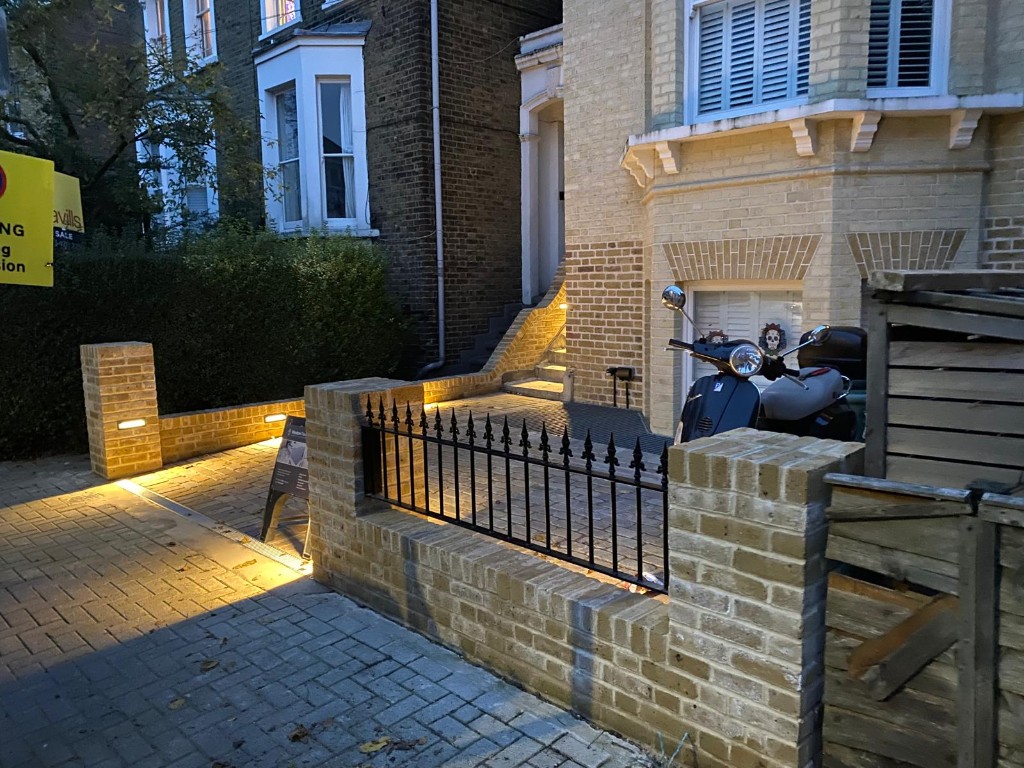 Buff brick walls and pillars with integrated warm recessed lighting beside block paving at dusk