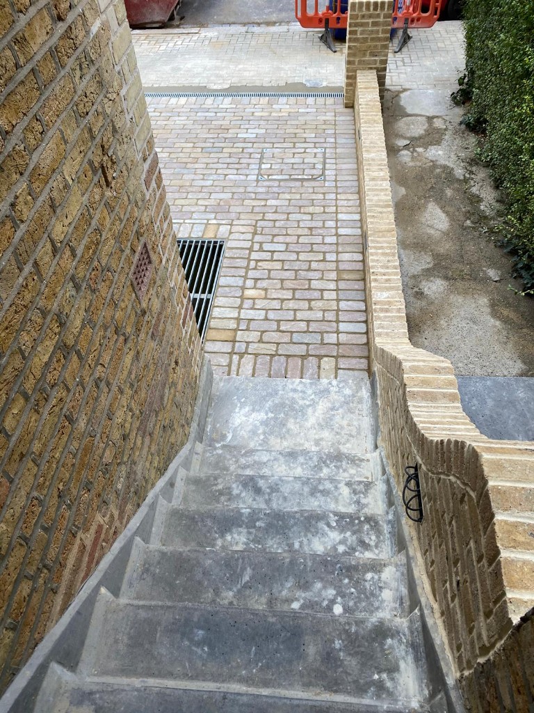 Concrete stair flight descending to buff paved landing beside a stepped yellow brick retaining wall