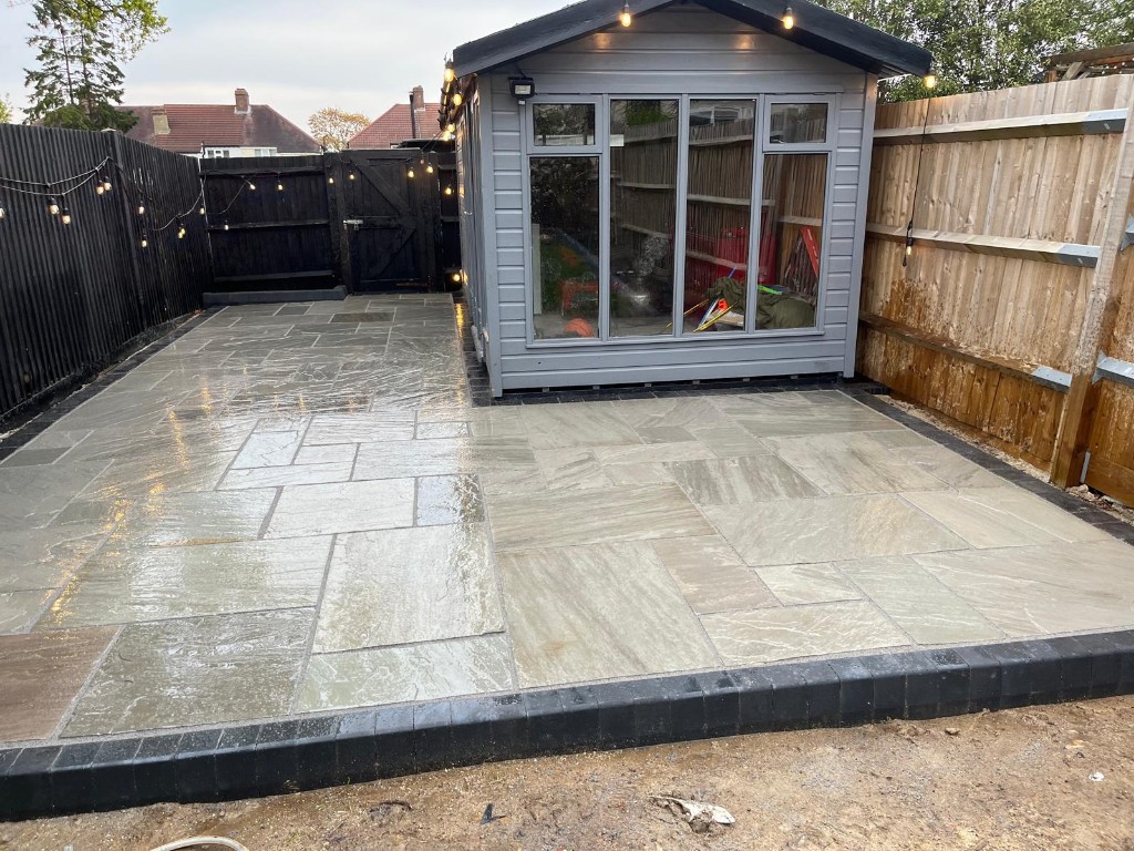 Wide natural stone patio with garden building and warm festoon lighting along the fence