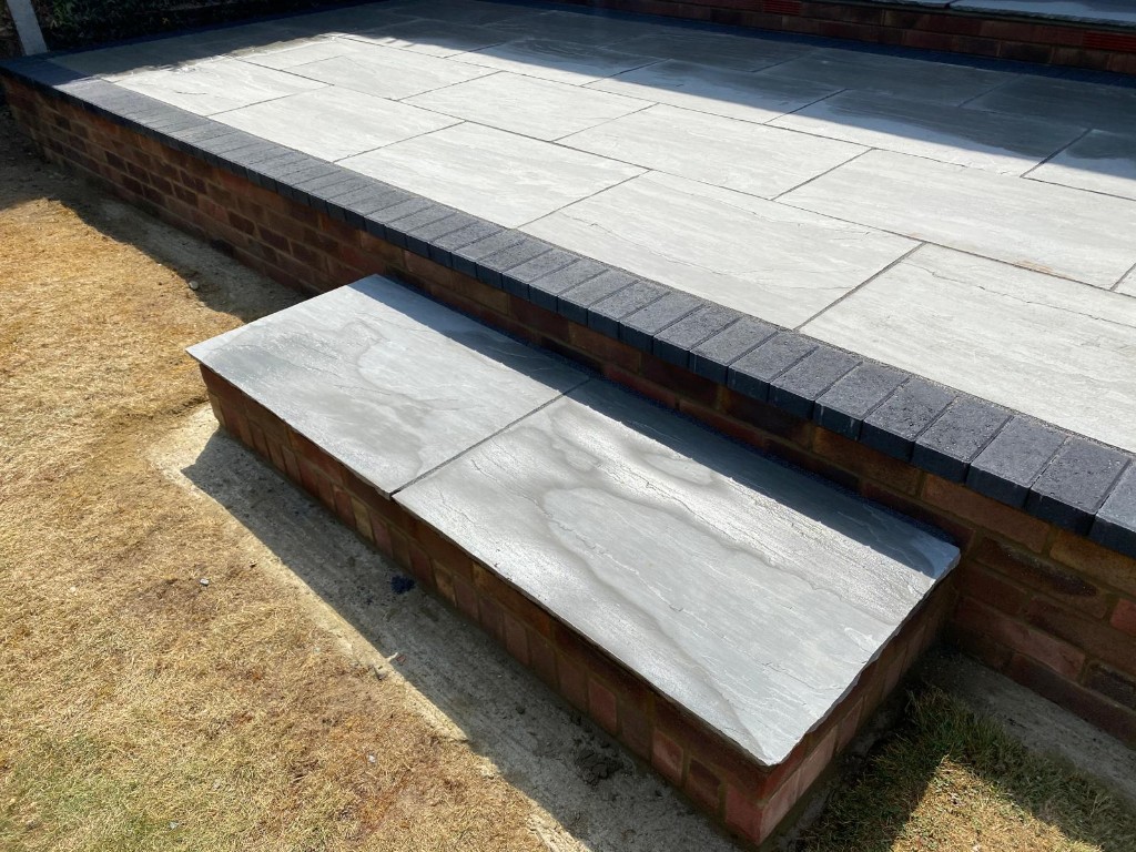 Raised light stone patio with dark block border and wide brick-faced step to lawn