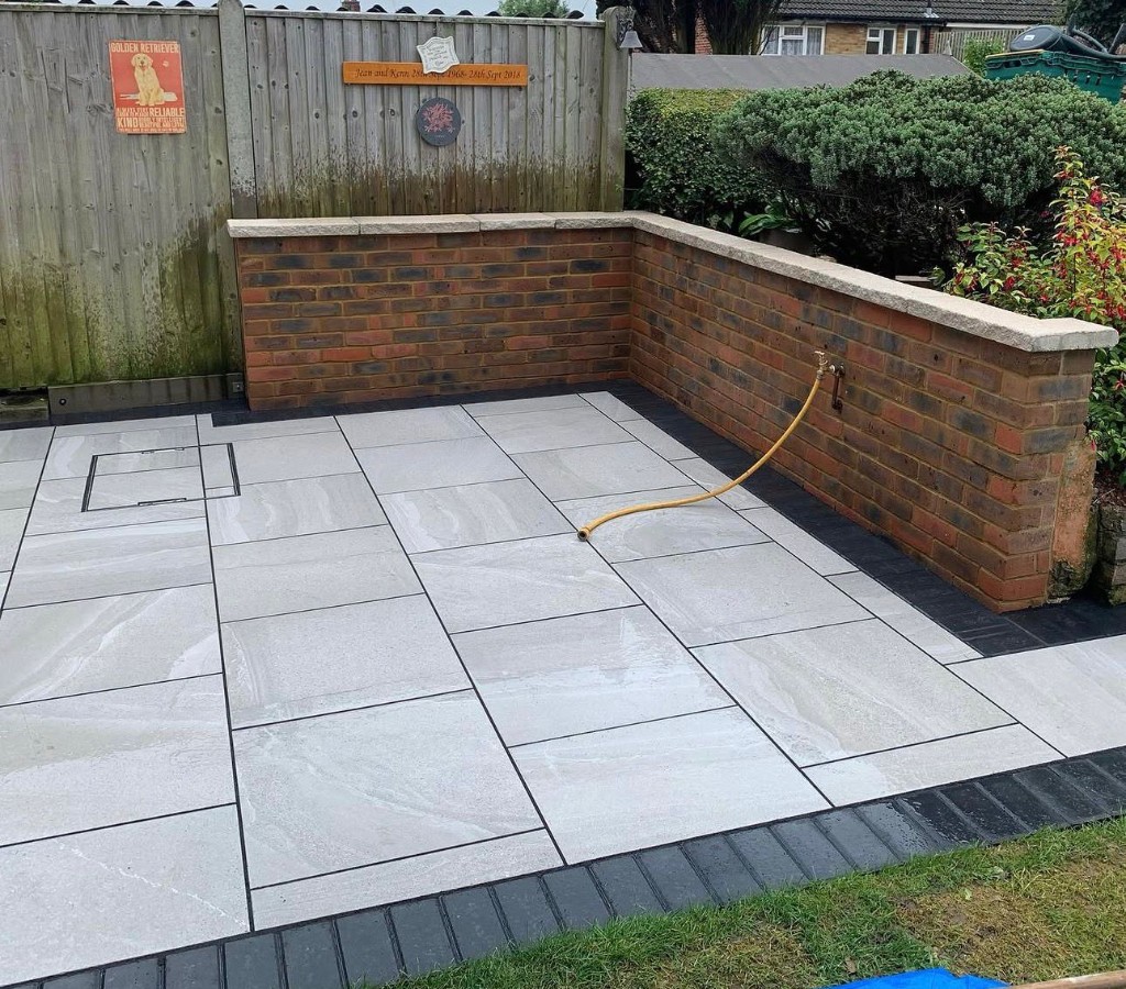 Light grey stone patio with dark joints and border, low brick wall with coping and paved-in utility cover