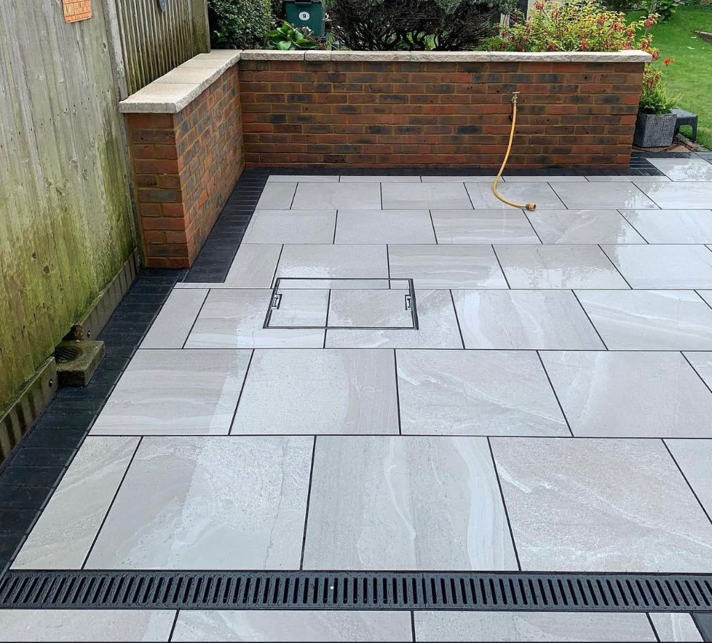 Contemporary grey patio with dark grout, block border, recessed utility cover and linear drainage channel