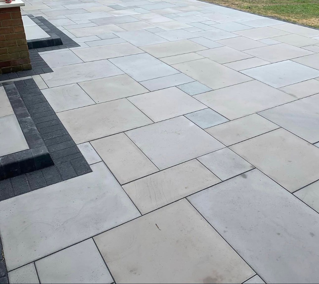 Light grey modular porcelain-style paving with charcoal border, shallow steps and lawn edge by a brick wall